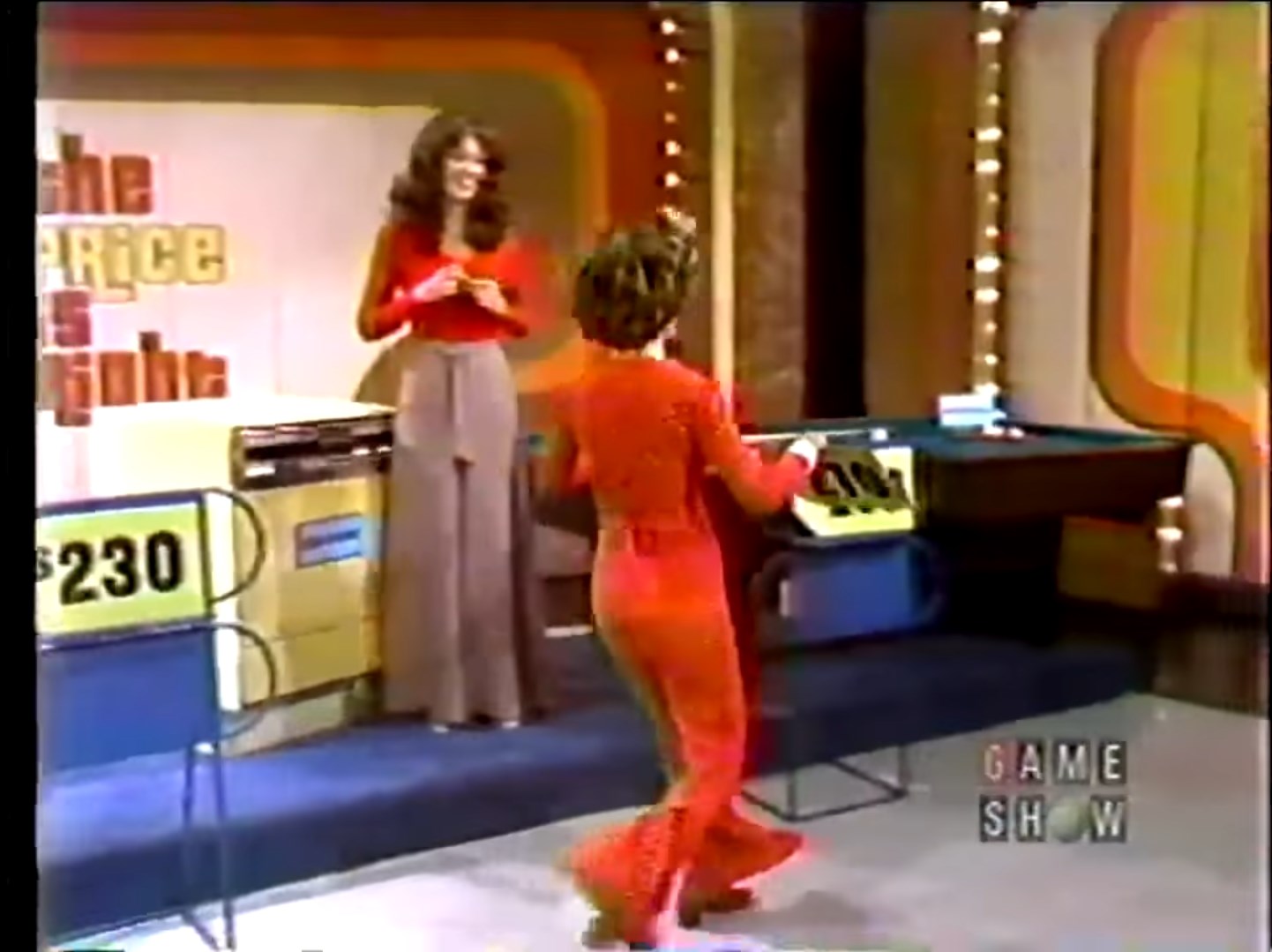 A contestant in a red outfit moves towards a display board showing the price tag of 0, while a host in a red top and gray pants smiles in the background, set onstage at The Price is Right in 1974.