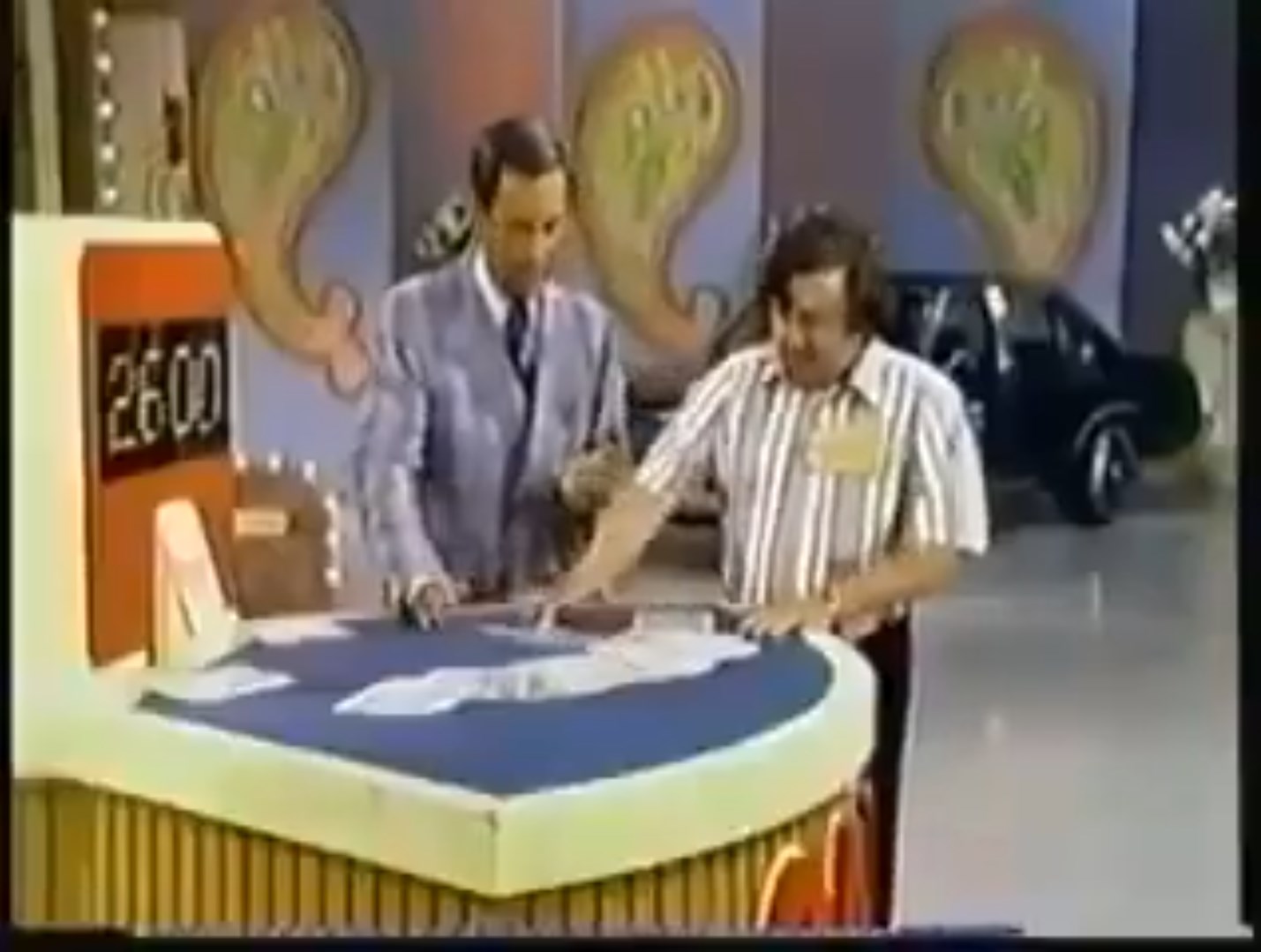 A game show scene featuring a host and a contestant at a card game table, with a car visible in the background. The host, dressed in a suit, appears to explain the game rules while the contestant looks engaged.