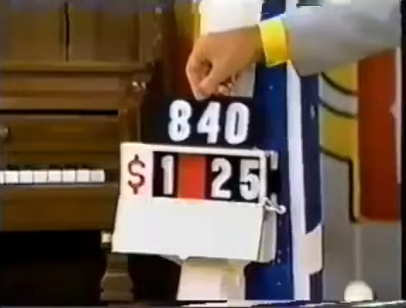 A hand holding a game board displaying the numbers 840 and a price of _25 in a colorful setting with a piano in the background.