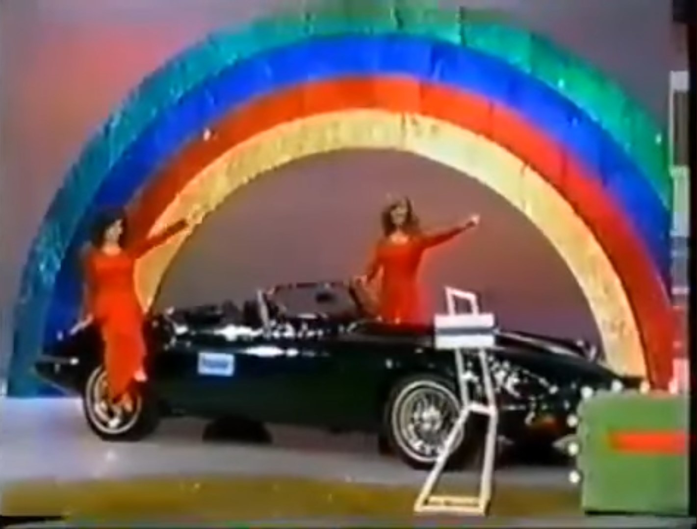 A colorful stage display with a rainbow backdrop featuring two women in red dresses gesturing towards a 1974 black convertible Jaguar XKE.