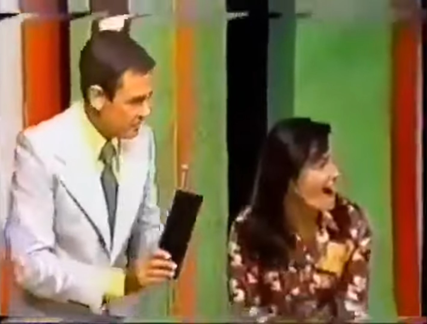 A man in a suit holds a black object while a woman beside him laughs joyfully, both appearing on a colorful game show set.