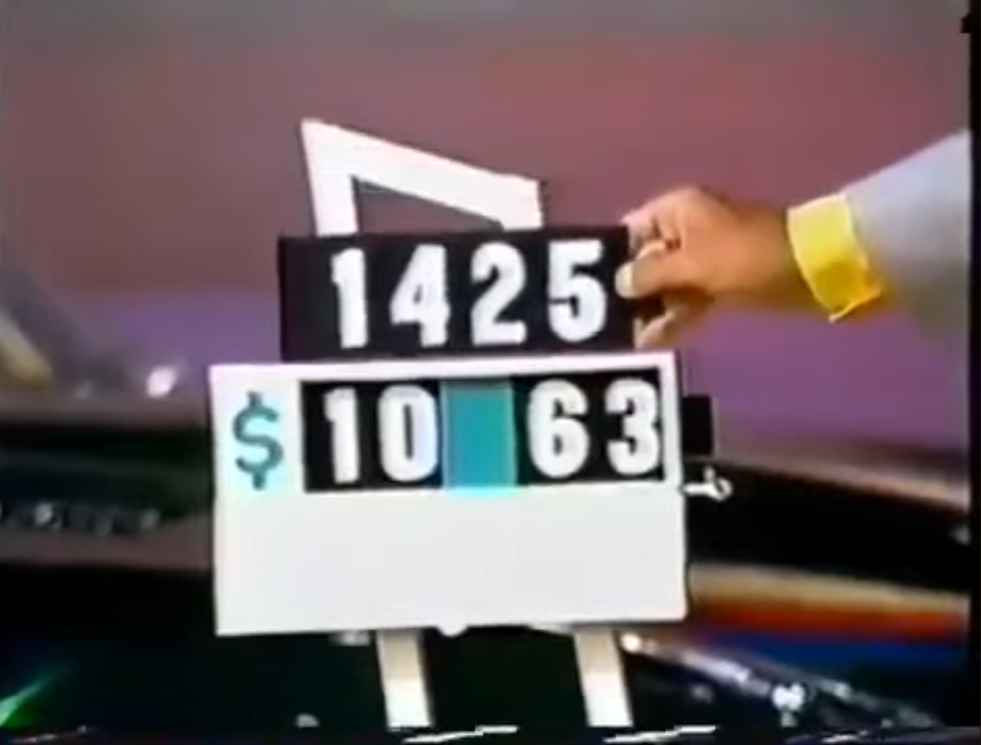 A person holding a game board displaying numbers 1425 and the dollar amount ,_63 during a game show.