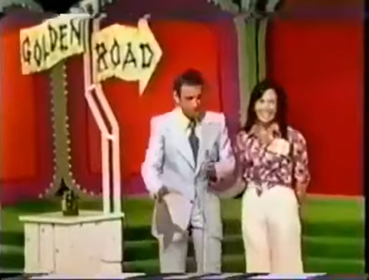 Bob Barker and a female contestant stand in front of a sign pointing to 'Golden Road', set against a colorful background, with a small jar visible on a pedestal.