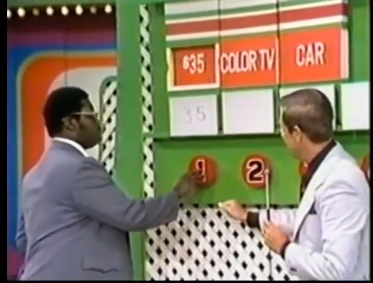 A contestant in a gray suit selects a number on a colorful game board during a pricing game on The Price Is Right, while the host observes.