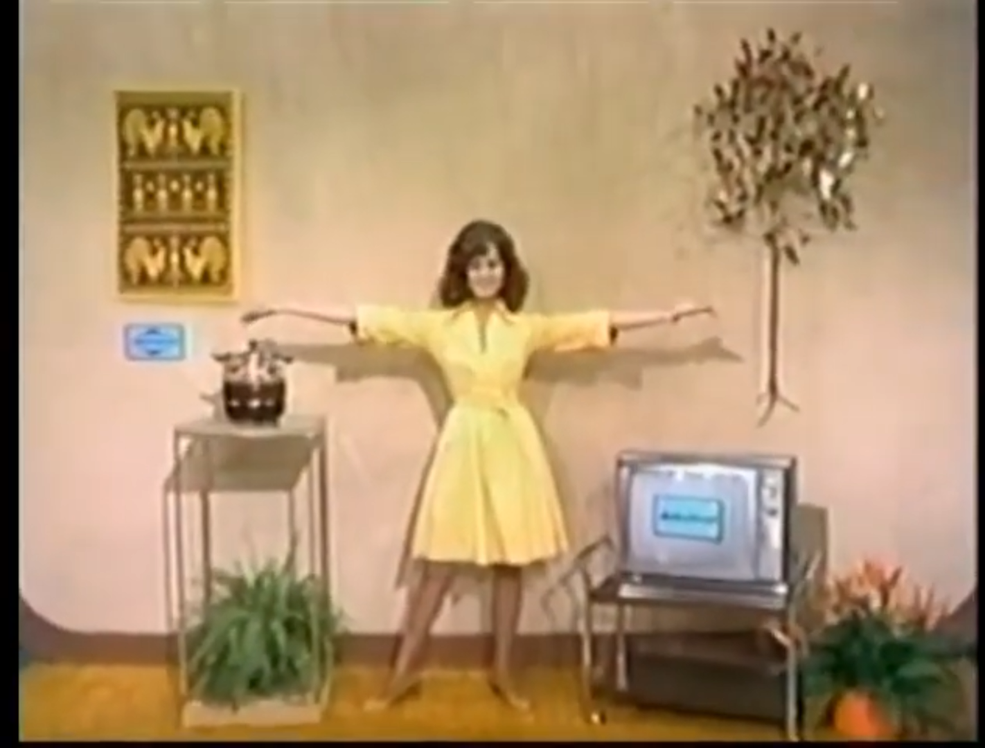 A woman in a yellow dress stands with arms outstretched next to a silver television, a potted plant, and a table with a beverage dispenser, against a beige wall with decorative art.