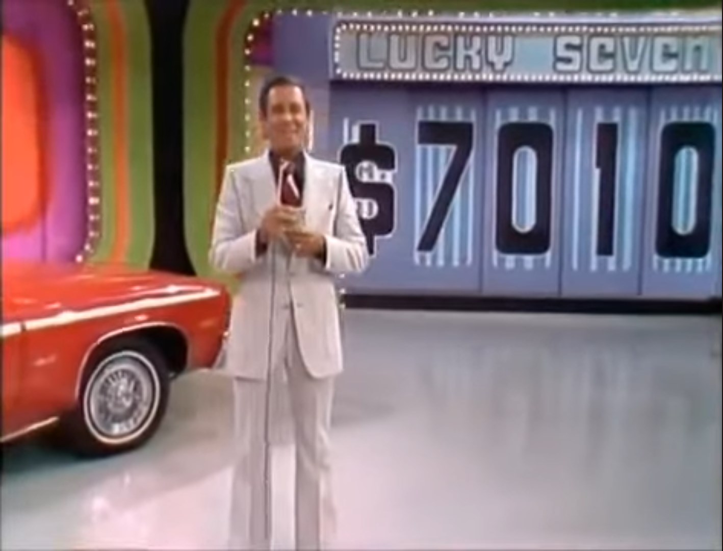 A host stands on stage with a microphone, presenting the game 'Lucky Seven' while a colorful backdrop and a red car worth ,010 are visible.