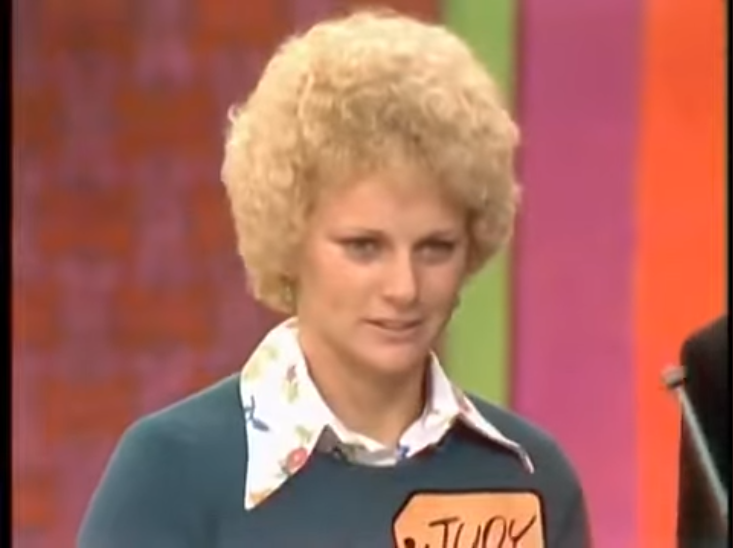 A contestant named Judy on a game show, looking thoughtfully while preparing to play the poker game, wearing a green sweater with a floral collared shirt.