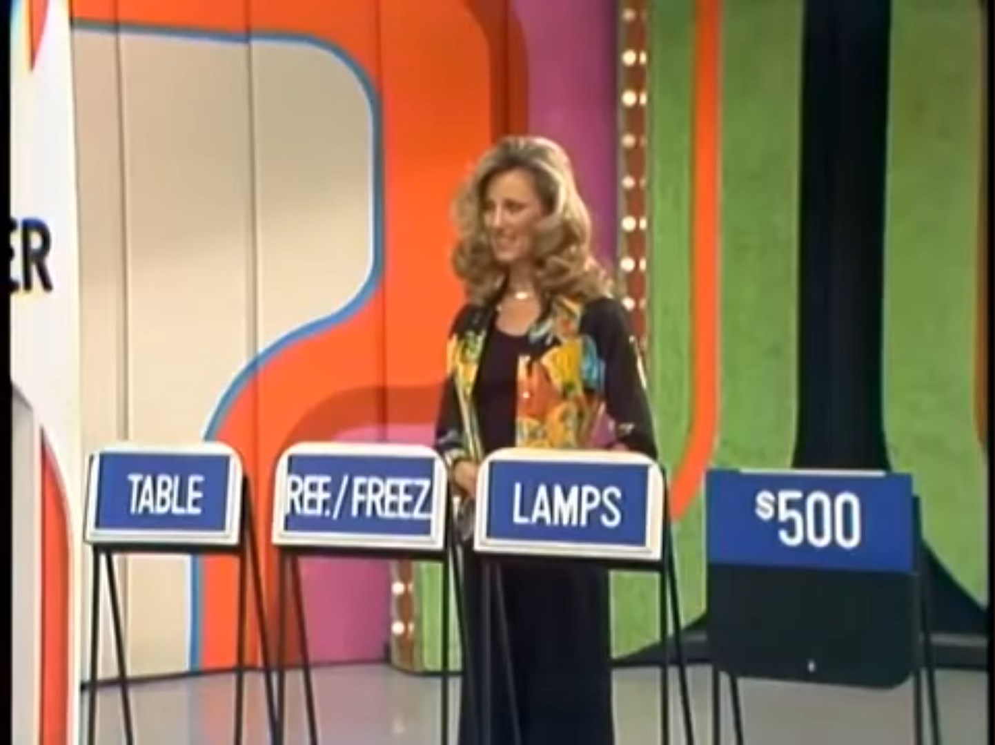 A game show contestant stands in front of prize displays, which include a table, refrigerator/freezer, and lamps, with a dollar amount of 0 visible.