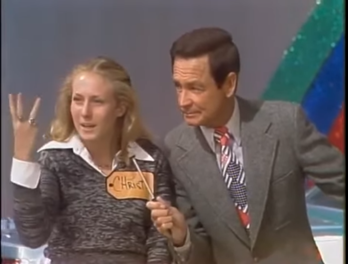A young woman with long hair, wearing a sweater and holding up three fingers, speaks with a male host wearing a suit and tie, both are engaged in a game show setting.
