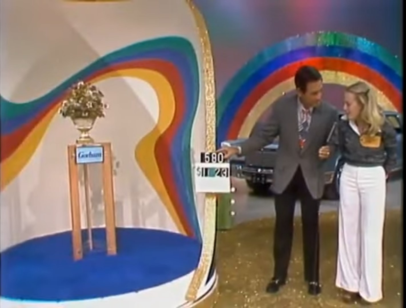 A game show host and a female contestant interacting with a prize display featuring a floral arrangement, with a colorful backdrop of rainbow stripes.