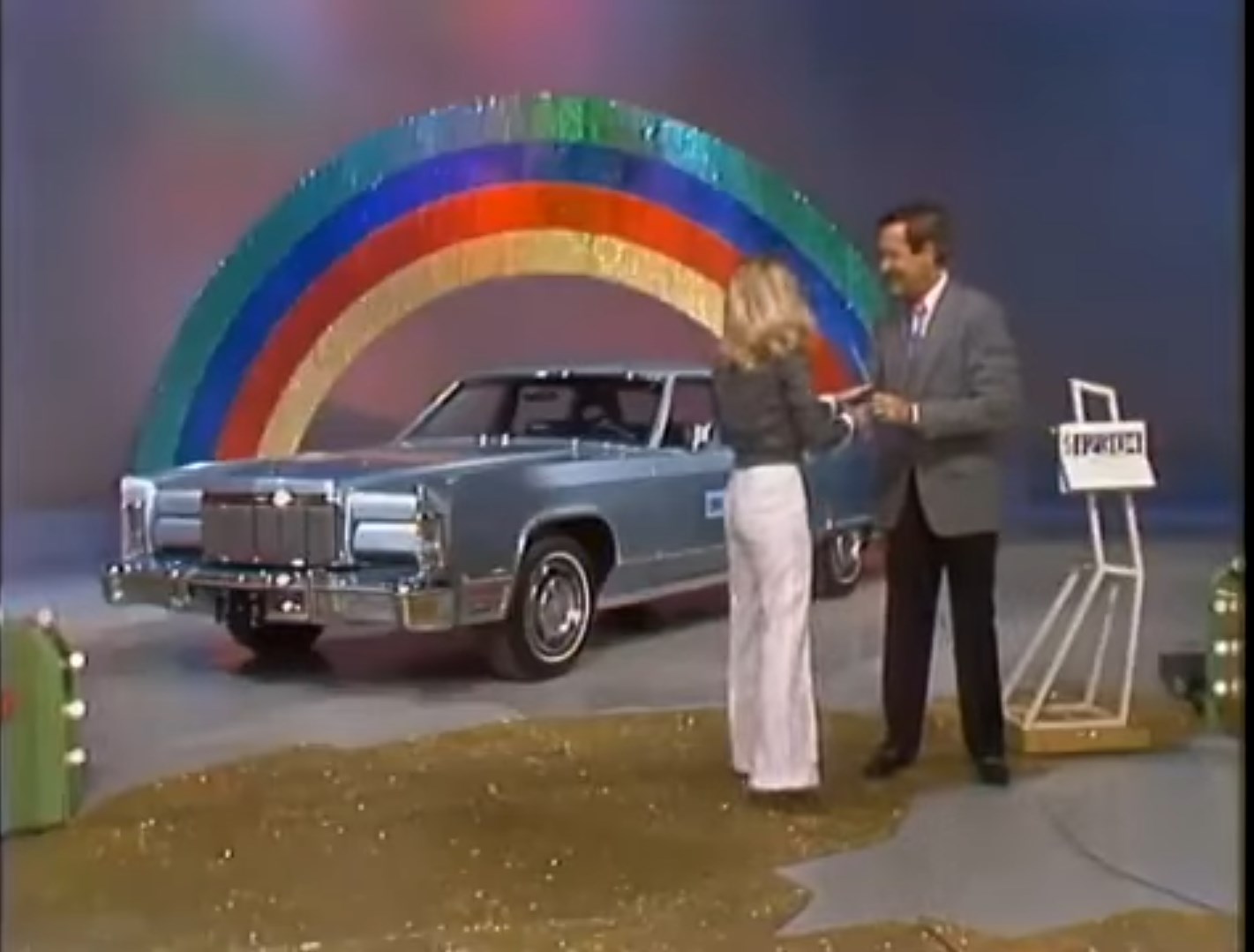 A contestant celebrates with the host in front of a classic car and a colorful rainbow backdrop on the set of a game show.