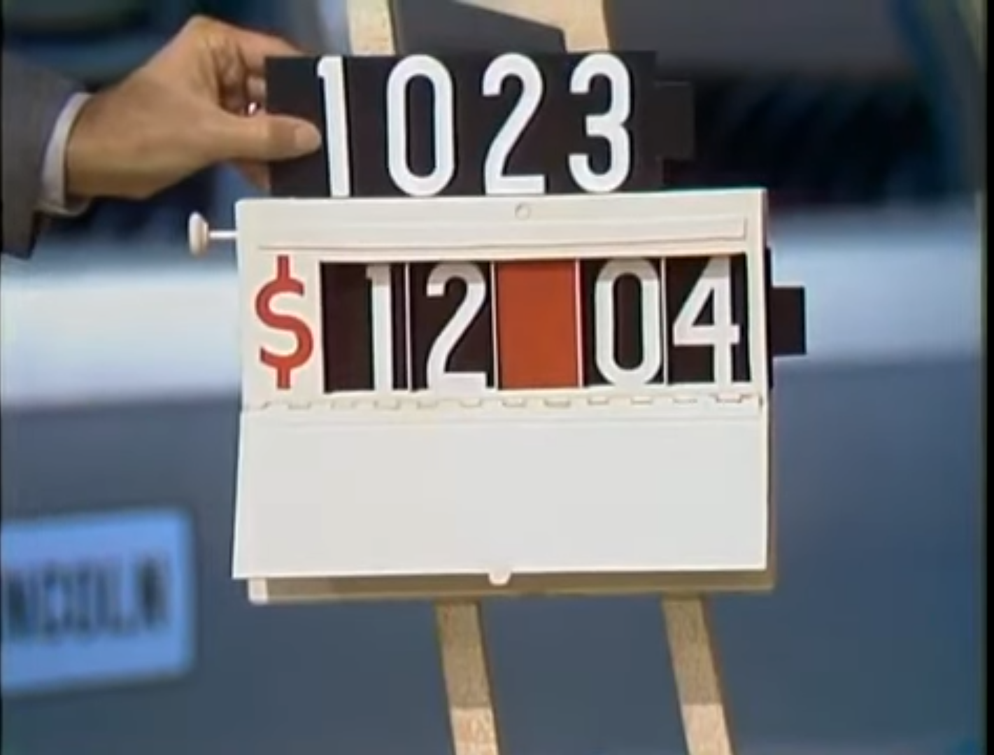 A scoreboard displaying the price of ,_04 with a hand adjusting the digits. The top row shows the number 1023.