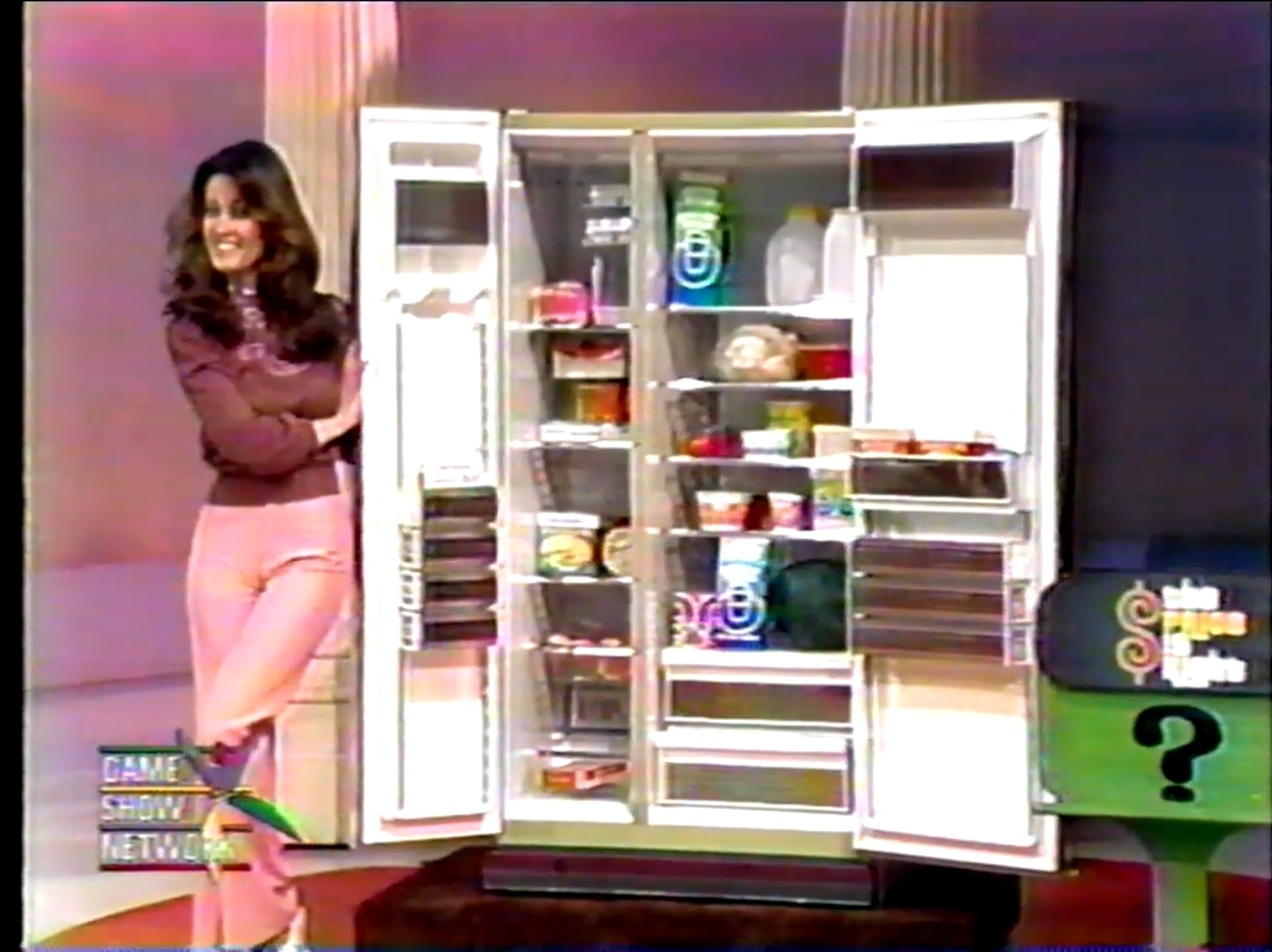 A model stands beside an open refrigerator filled with various food items, showcasing excitement during a game show segment.
