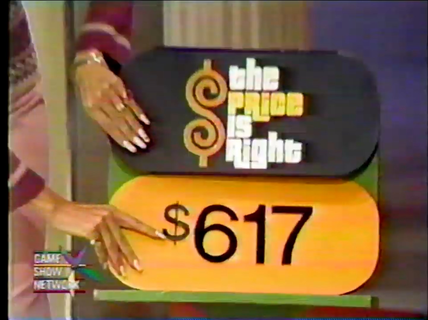 A game show host points to the price tag displaying 7 for the game 'The Price is Right'.