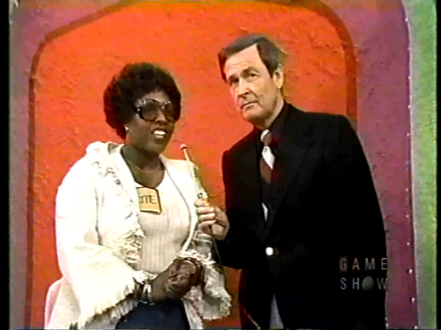 A contestant wearing sunglasses and a name tag holds a microphone while speaking with the host against a vibrant orange background.