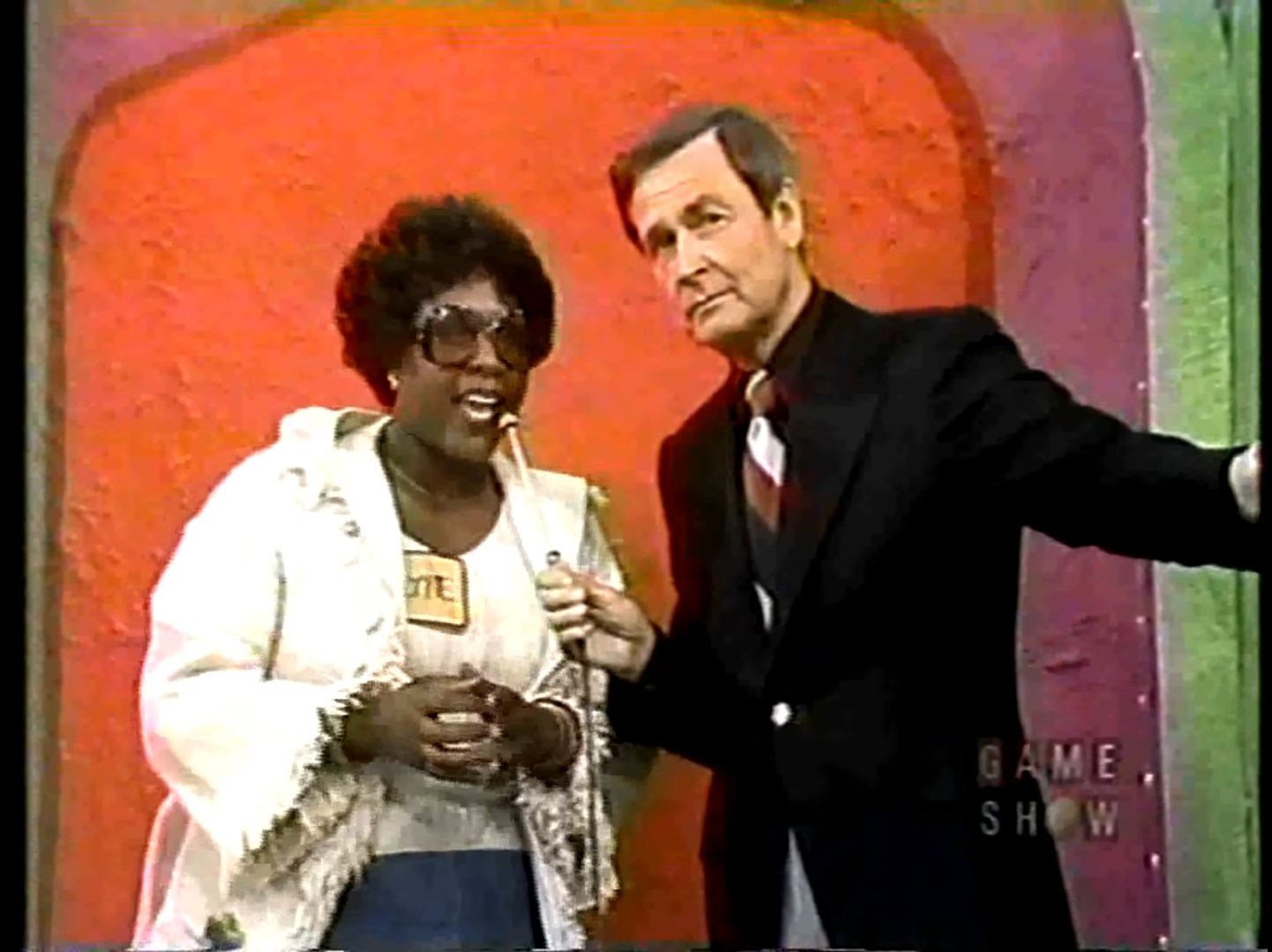 A contestant enthusiastically speaks into a microphone while standing next to Bob Barker in a colorful set background.