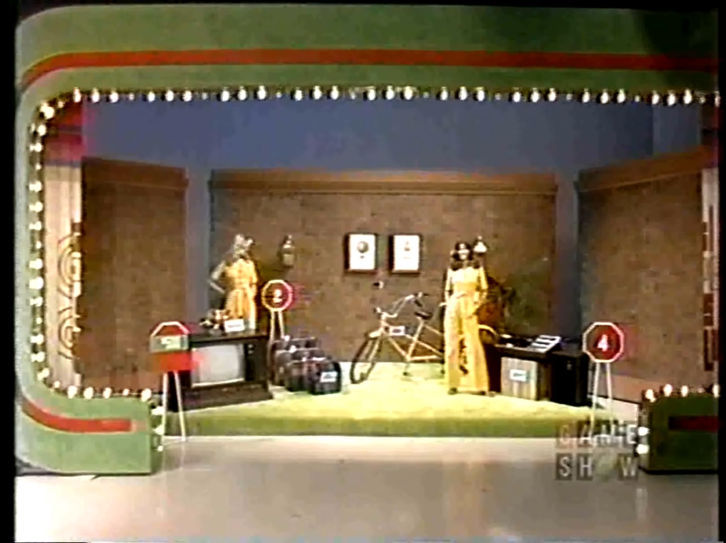 A game show set for the 'Danger Price' game featuring prizes including a color TV, luggage, a tandem bike, and a stereo, with two models in yellow dresses.