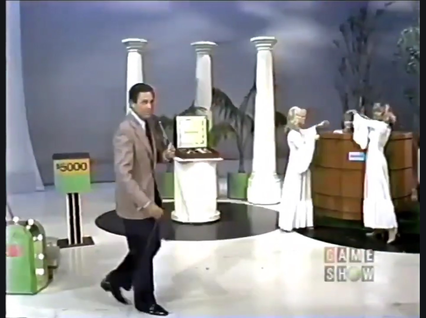 A game show host walks on stage with two models presenting prizes in a set designed with columns and palm trees.