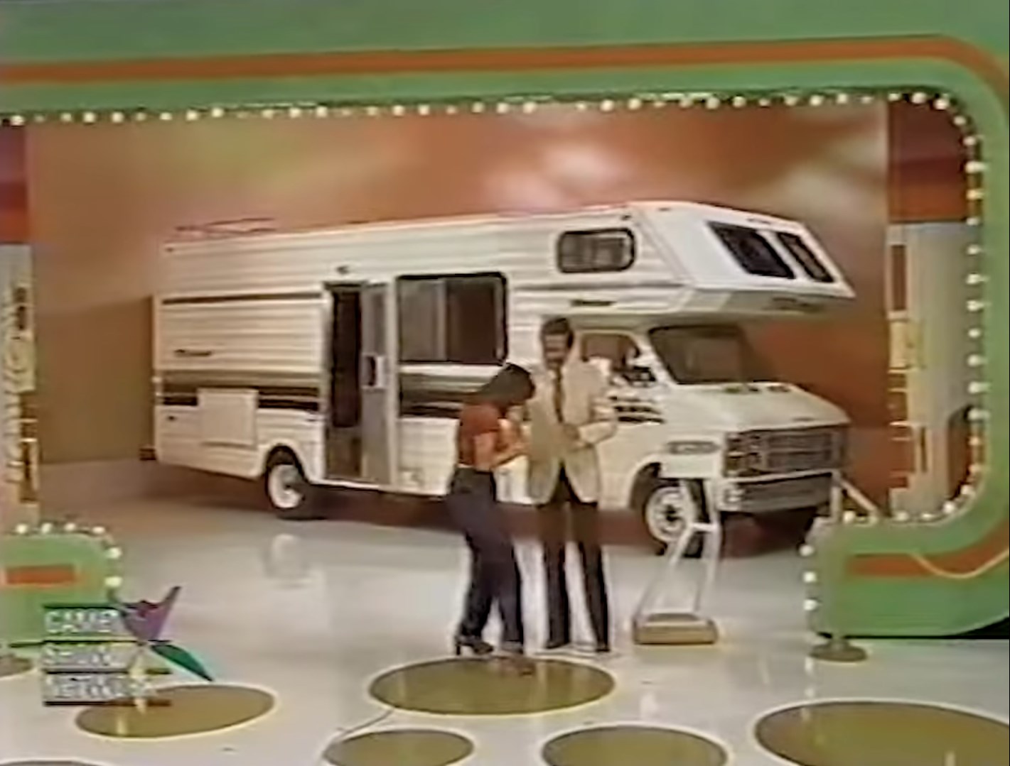 A contestant celebrates with the host in front of a large motorhome on set during a game show.