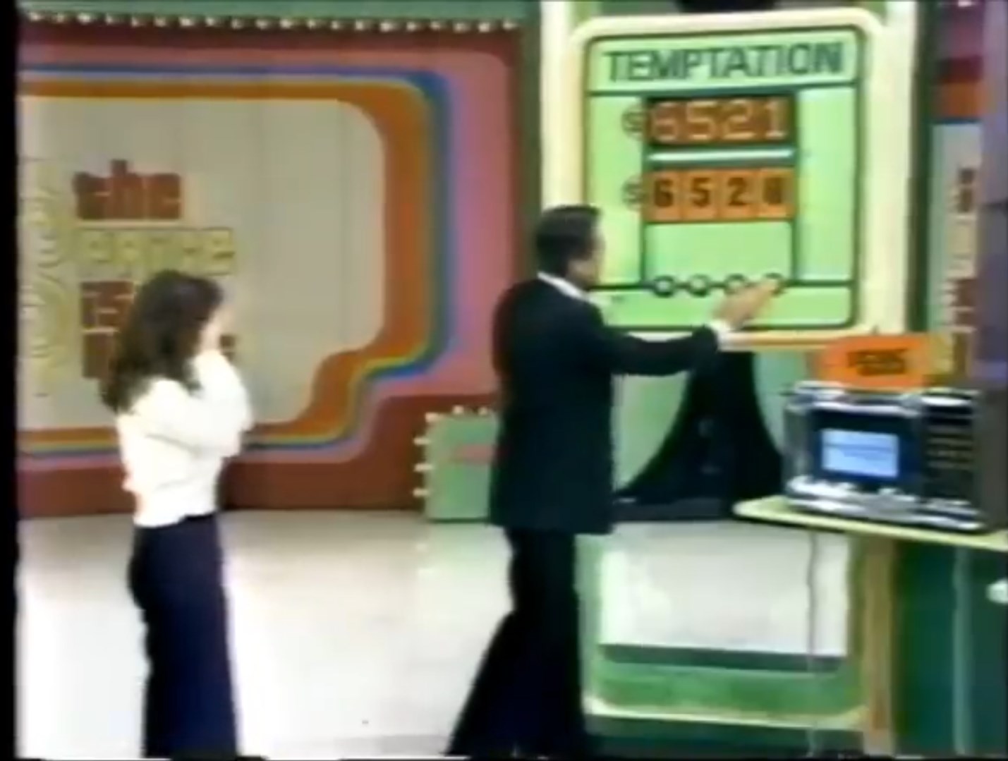 A contestant watches as the host points to the game board labeled 'TEMPTATION', displaying the numbers 21 and 20 during a game show.