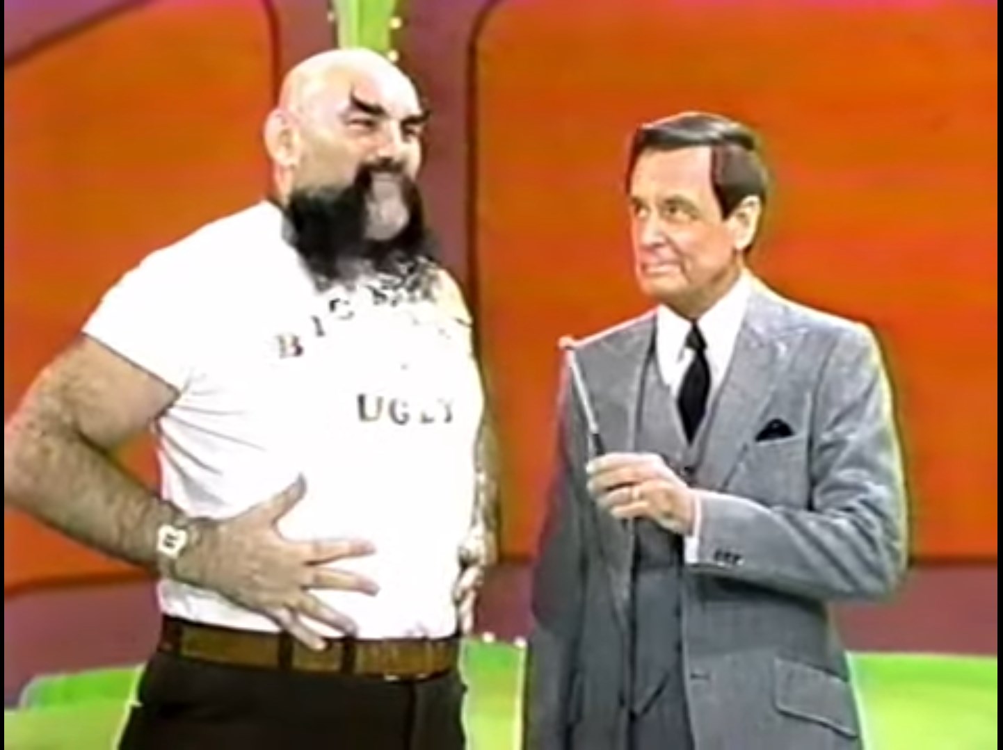 A humorous scene from a game show featuring Ox Baker in a 'Bigger is Ugly' t-shirt and a suited Bob Barker looking nervous.