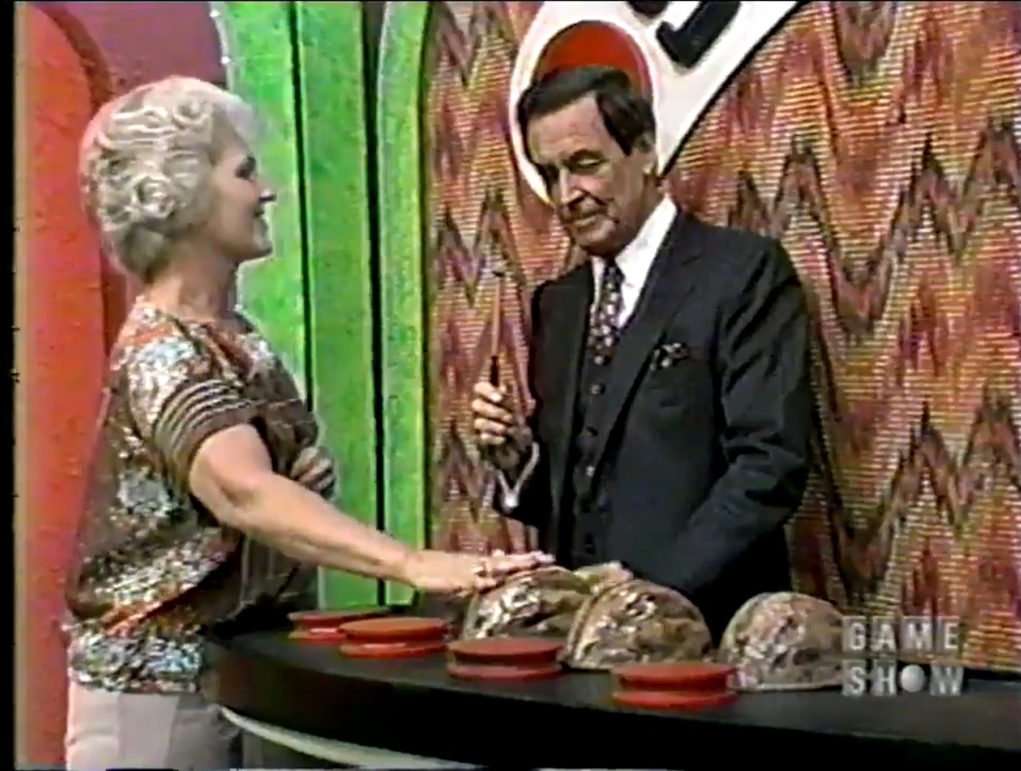 A game show host presents the Shell Game with four shells in front of a contestant, who is reaching towards the shells.