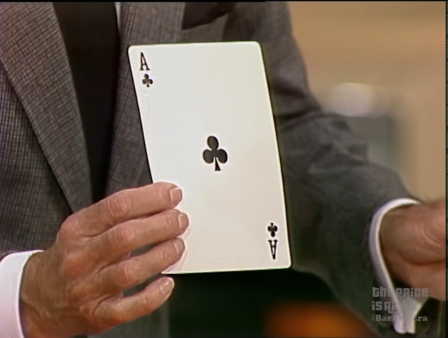 A contestant holds up an Ace of Clubs during a card game, showcasing the card's value.