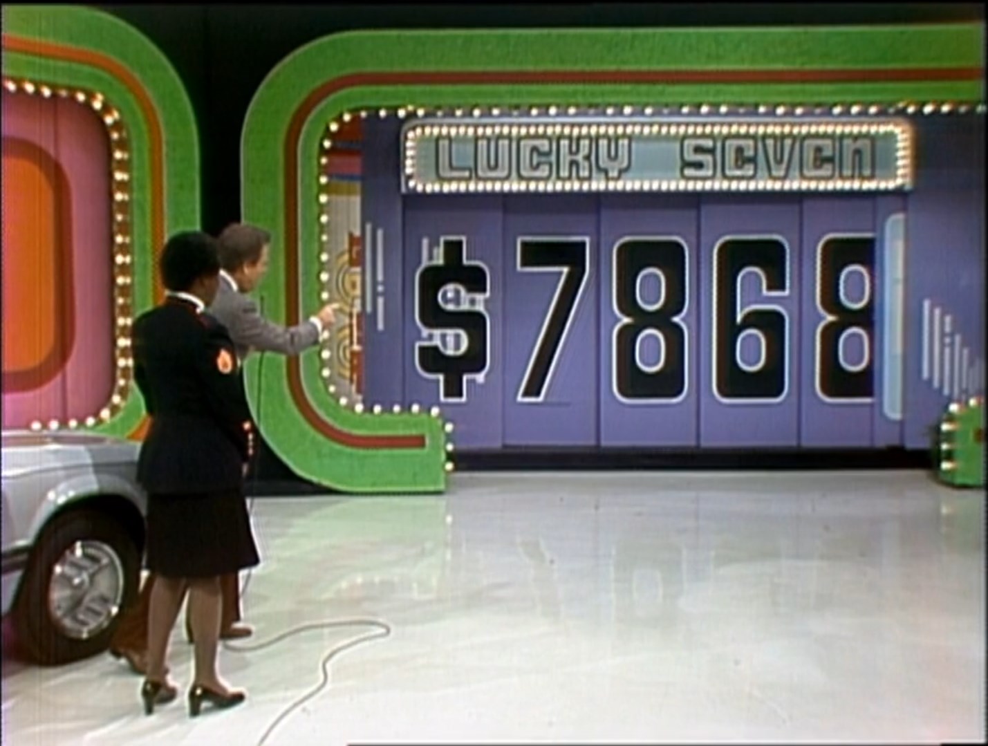A game show scene showing the host pointing at the price of a car displayed in the Lucky Seven game with the digits 68 visible on the board.