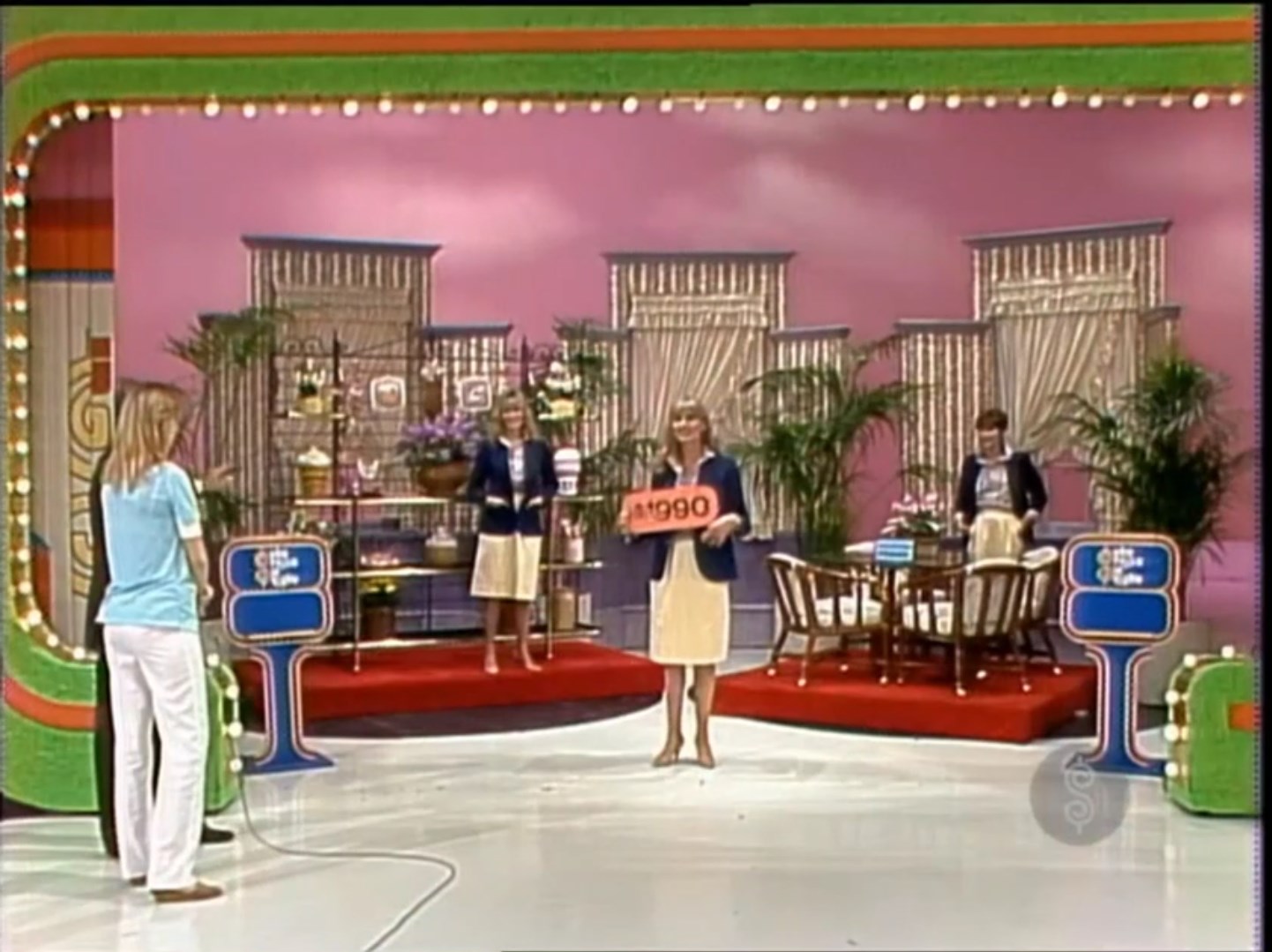 A game show stage featuring contestants and models, with a price tag of 0 displayed. The set is decorated with plants and curtains, and two models are posing next to a dinette set in a vibrant, pink backdrop.