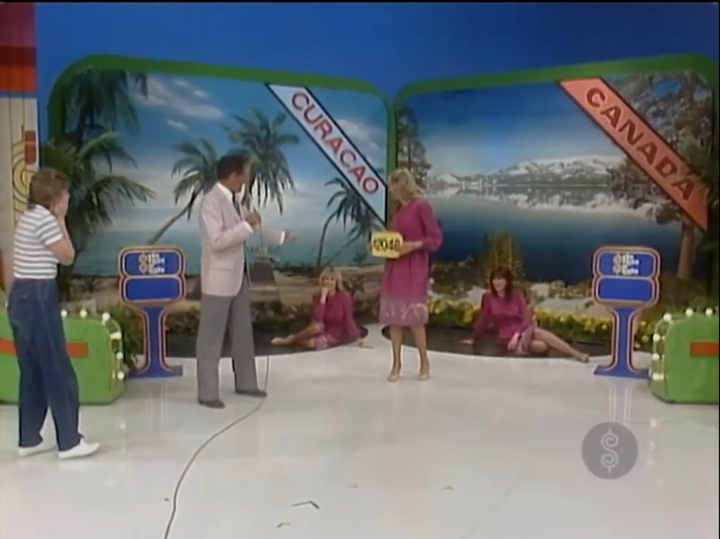 A game show host and a contestant discuss prizes while two models sit on the floor. The backdrop features scenes of Curaçao and Canada, with price tags displayed.