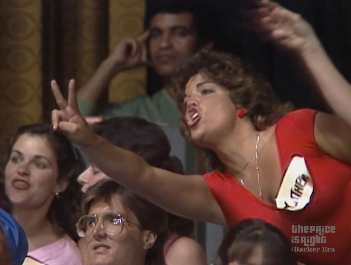 A passionate contestant in a red shirt enthusiastically gestures with two fingers while shouting in a game show audience.