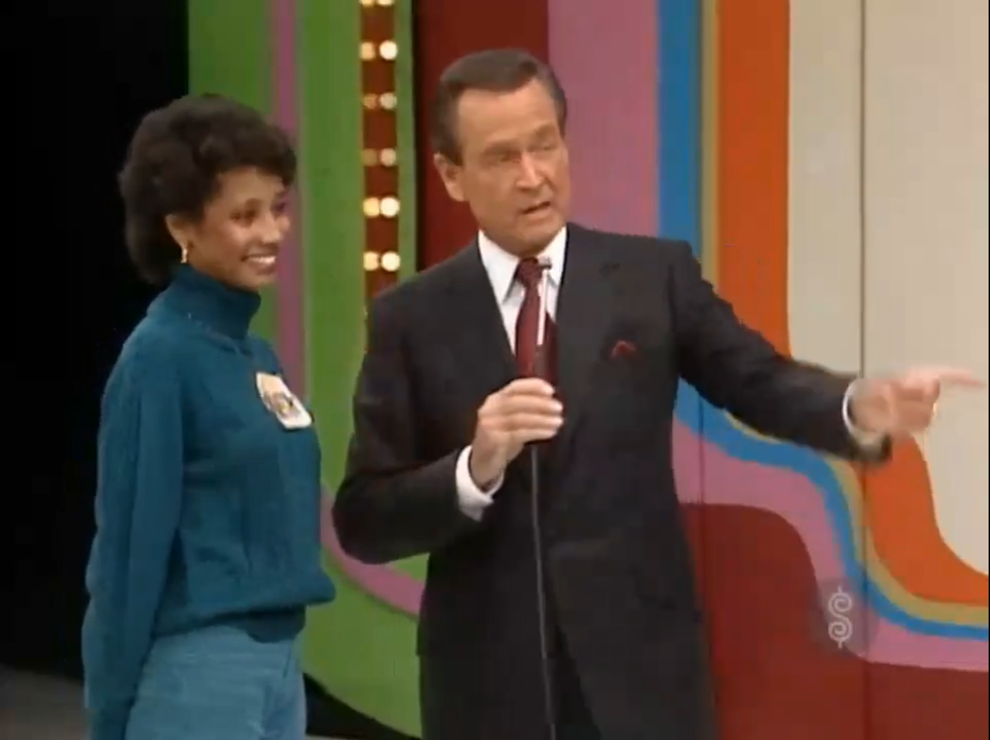 A host points while standing next to a contestant on a game show set, with colorful background elements.