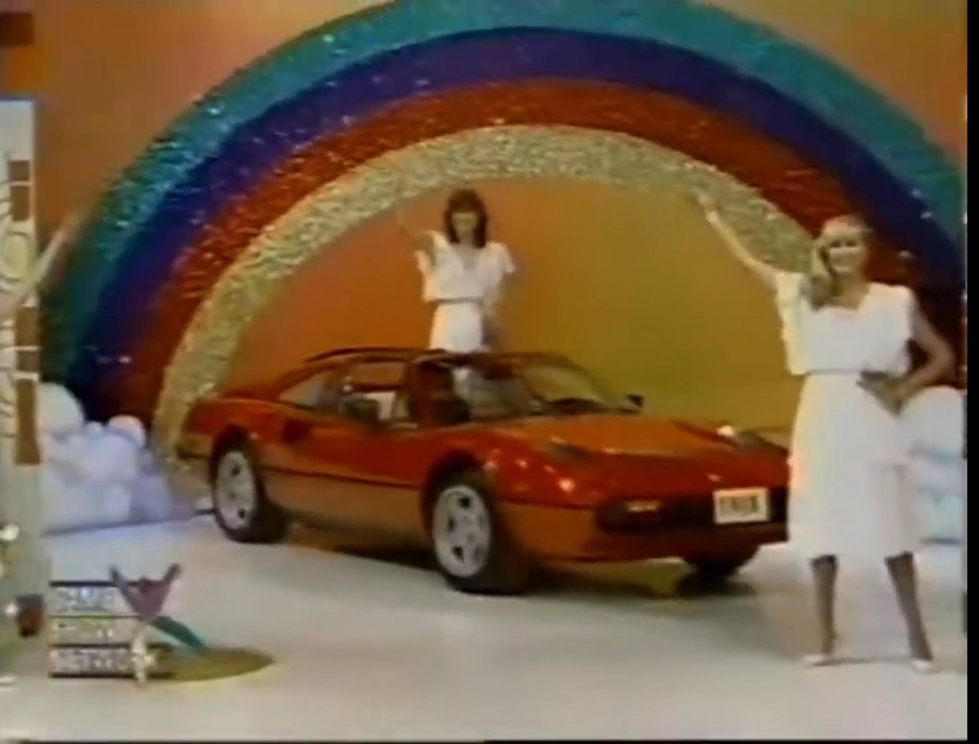 A bright stage featuring a red car under a rainbow backdrop, with two women in white dresses waving at the audience.