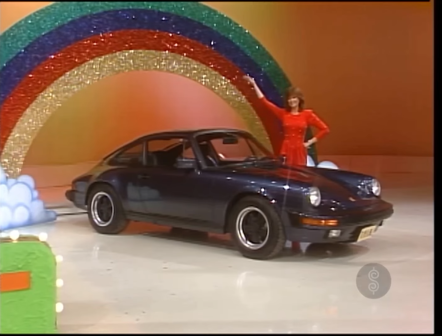 A contestant in a red dress stands next to a dark blue Porsche 911 Carrera under a sparkling rainbow backdrop on a game show set.