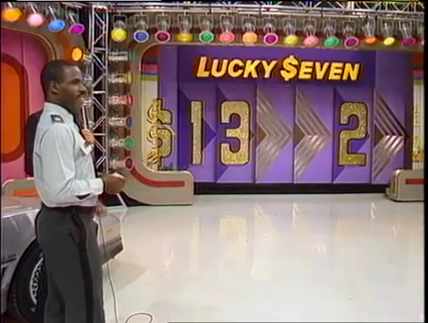 A game show set for 'Lucky Seven' featuring a host standing in front of colorful podiums displaying numbers 1, 3, and 2, with the show's name and dollar signs in bold lettering.