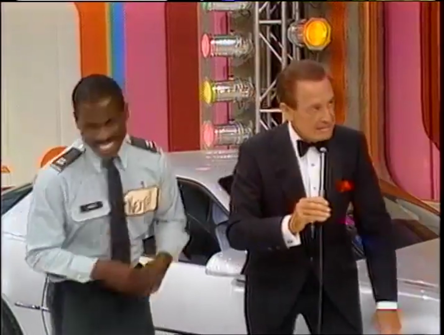 A smiling contestant in a military uniform stands beside a classic car while Bob Barker speaks into a microphone on stage.