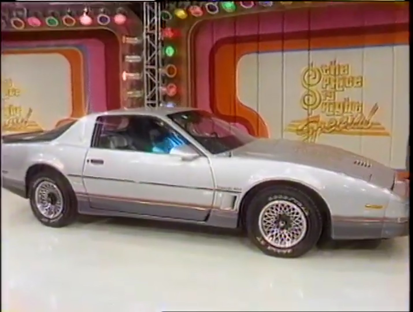 A silver Trans Am displayed on stage at The Price is Right in a 1986 primetime special, with colorful lights and a backdrop featuring the show's logo.