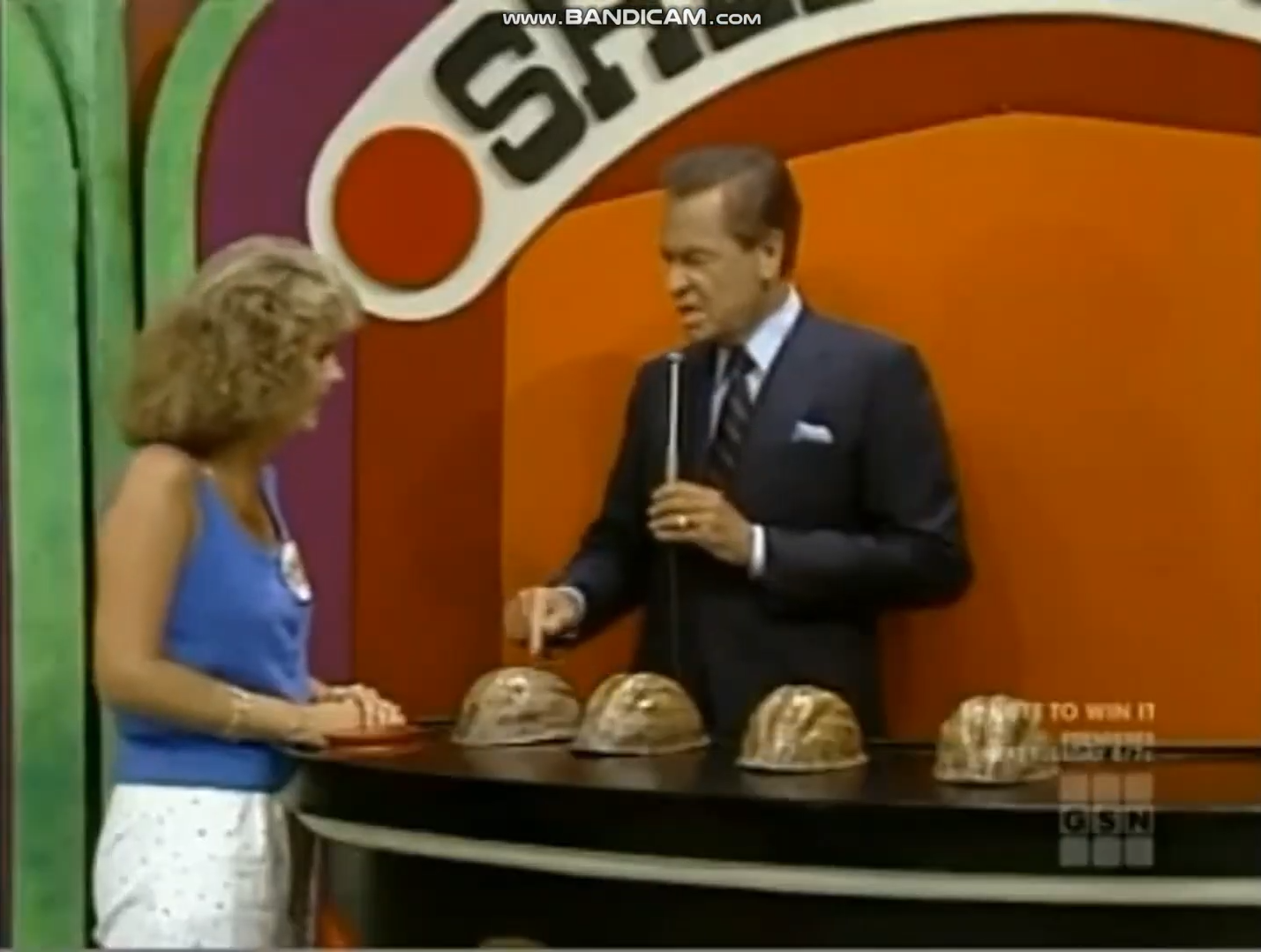 A host explains the Shell Game rules to a contestant, with four shells displayed on a game show set.