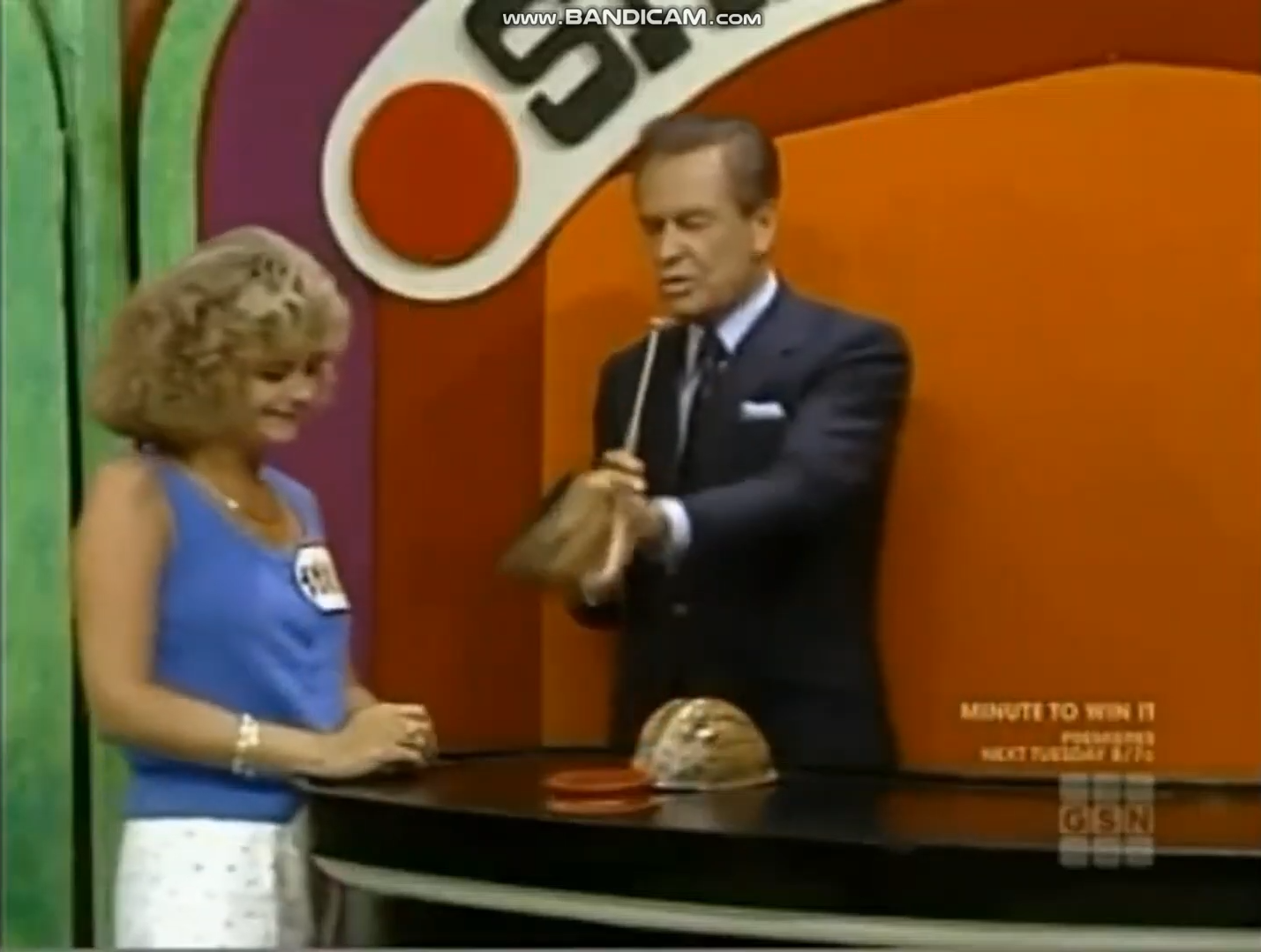 A female contestant smiles while the host demonstrates the Shell Game, holding a shell as they engage in a game show setting.