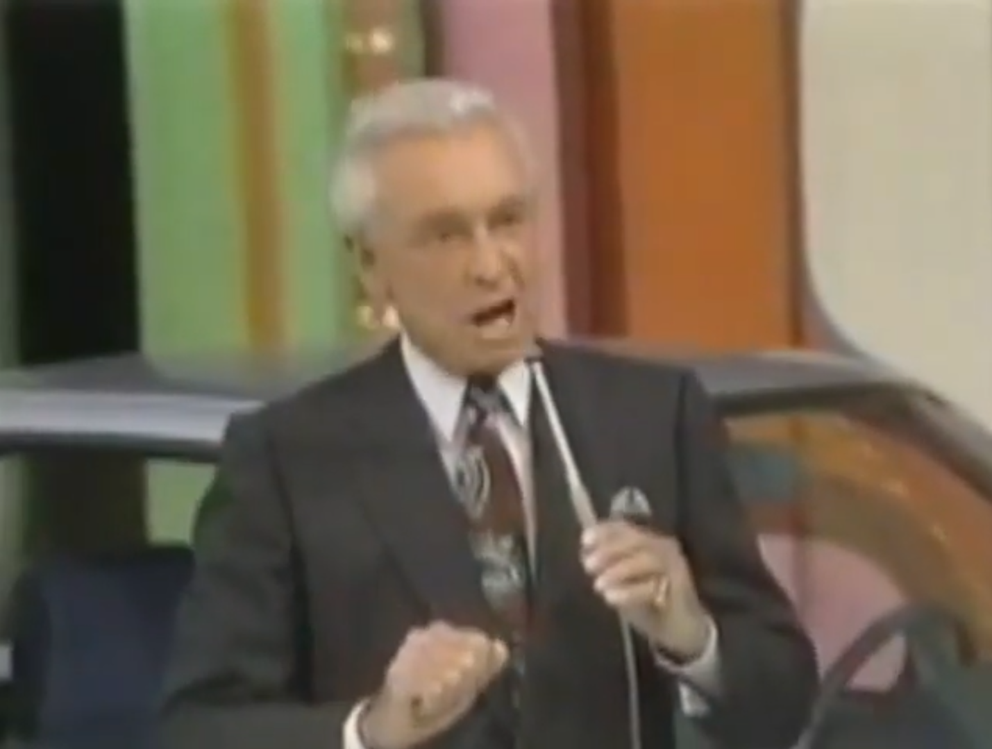 Bob Barker enthusiastically speaking into a microphone in front of a colorful backdrop, with a car partially visible behind him.