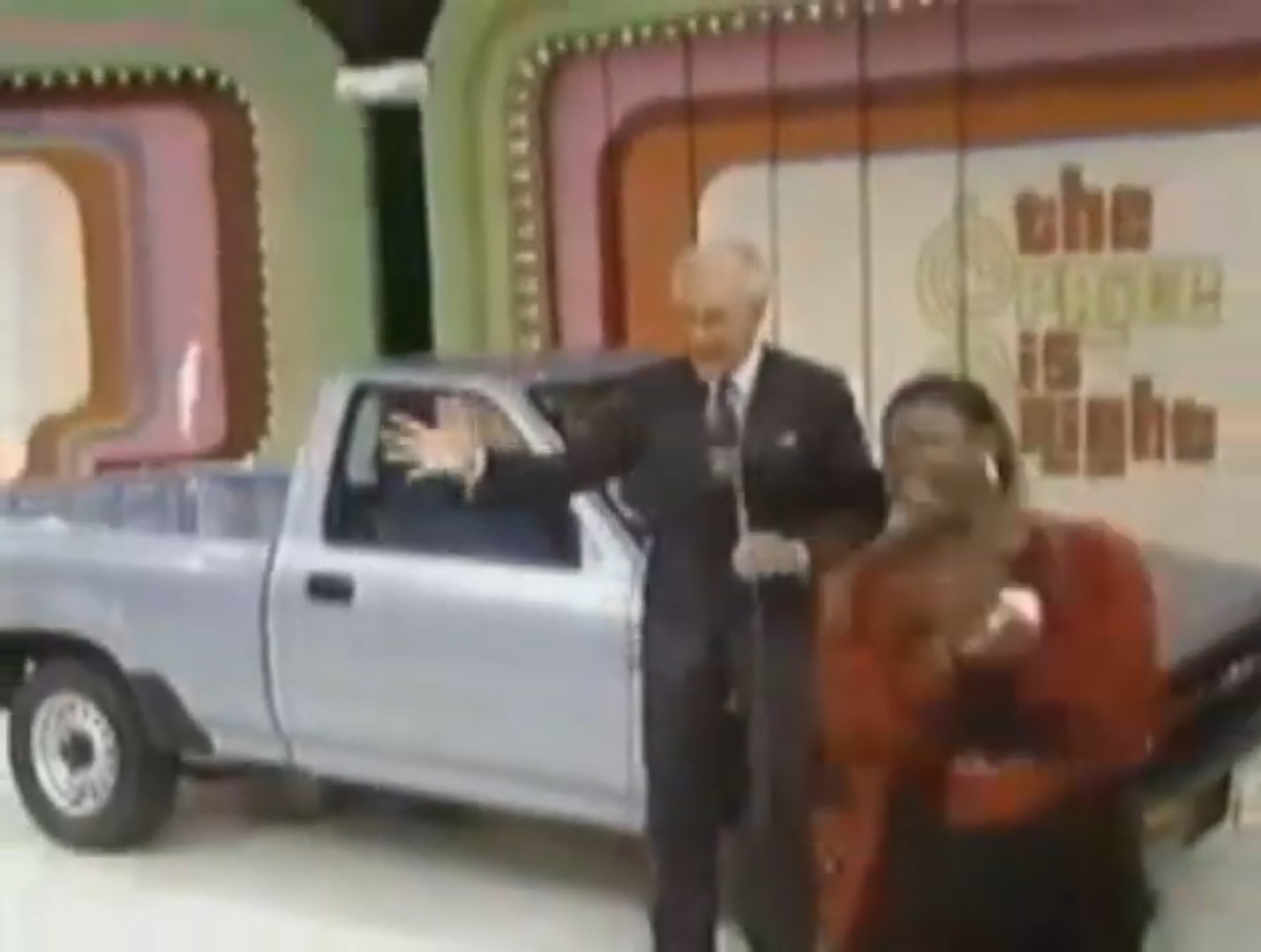 A game show host presents a contestant celebrating next to a silver pickup truck on stage.