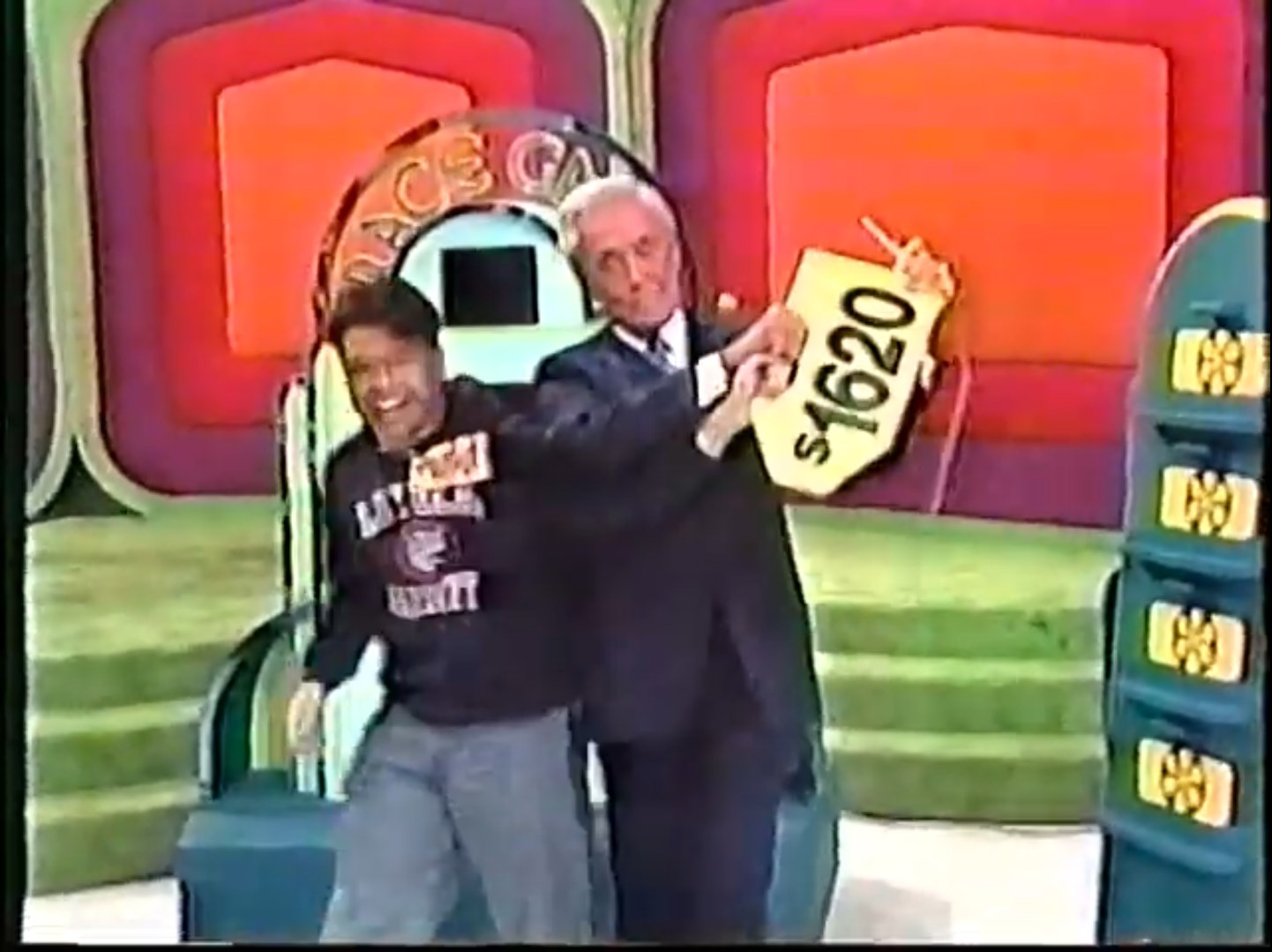 A contestant joyfully celebrates with a host on the set of The Price is Right, holding a price tag showing 20, with a colorful backdrop and game display in view.