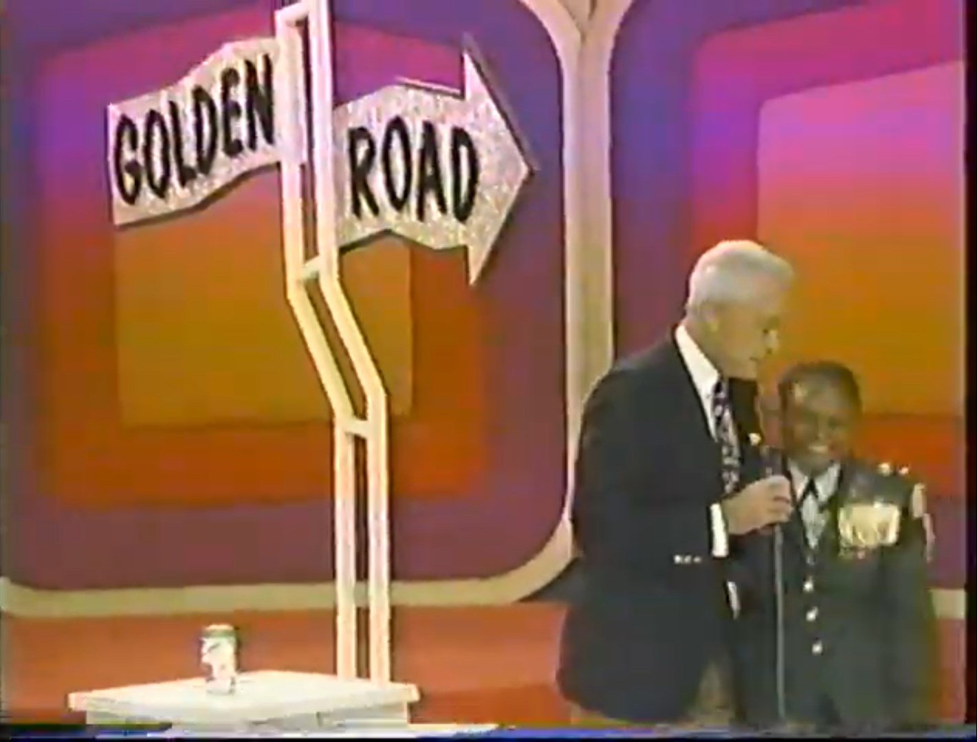 A television host interacts with a young contestant standing beside a sign indicating the 'Golden Road' game, set against a colorful backdrop.