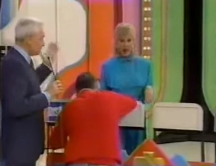 A scene from the Poker Game on a game show, featuring a host speaking into a microphone, a contestant in a red shirt interacting with a game table, and a female presenter in a blue outfit observing.