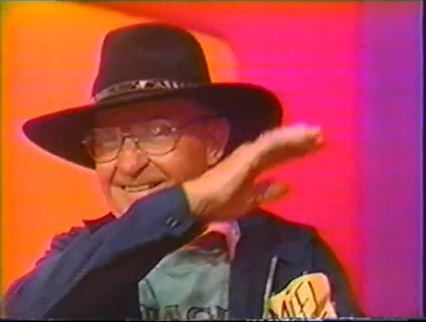 A contestant smiling and waving while wearing glasses and a hat, with colorful backgrounds.