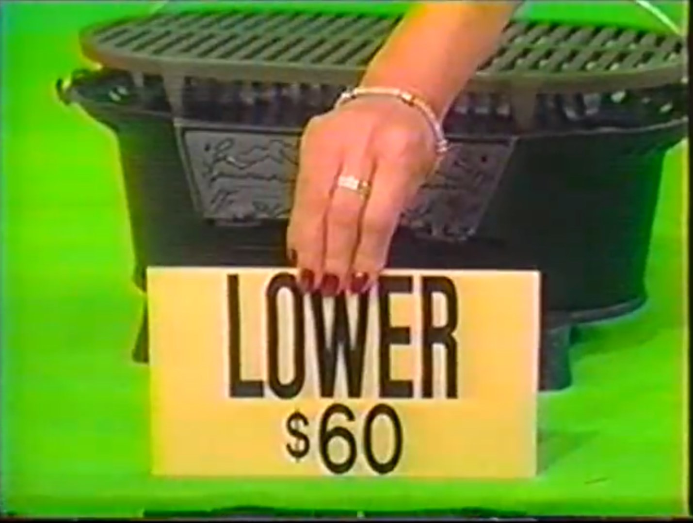A contestant holds up a sign saying 'LOWER ' next to a black grill on a colorful background.