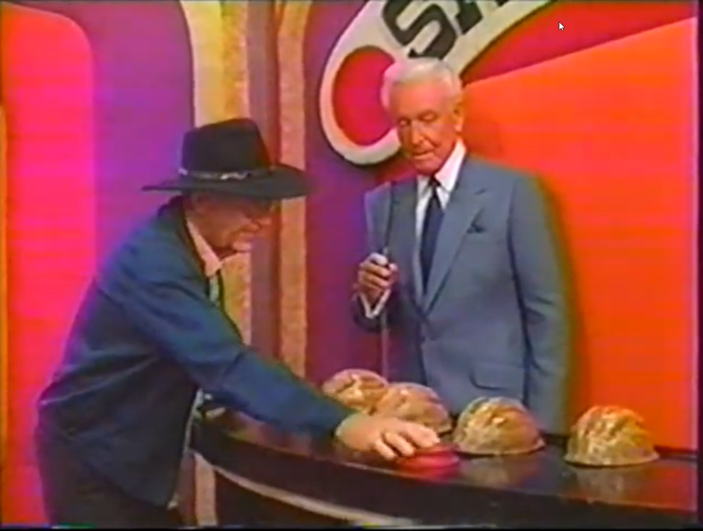 A contestant wearing a black hat is interacting with colorful shells on a game show set, while the host stands beside him holding a microphone.