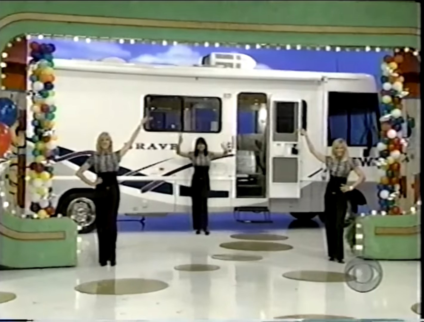 Three models presenting a travel trailer during a game show, with colorful decorations and stage lights in the background.