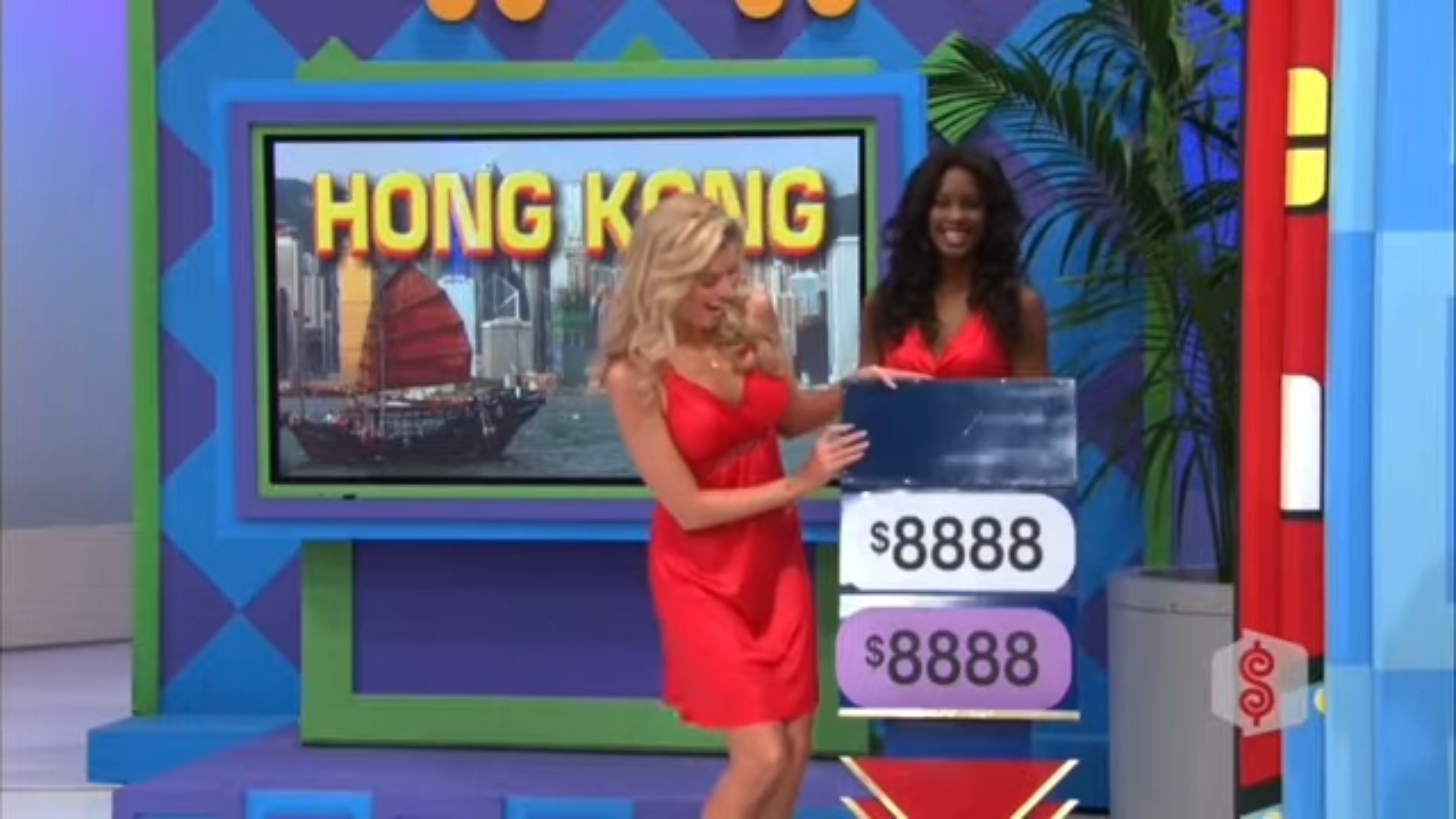 A model in a red dress holds up a blue board showing a price of 88 next to another contestant as the backdrop features an image of Hong Kong.