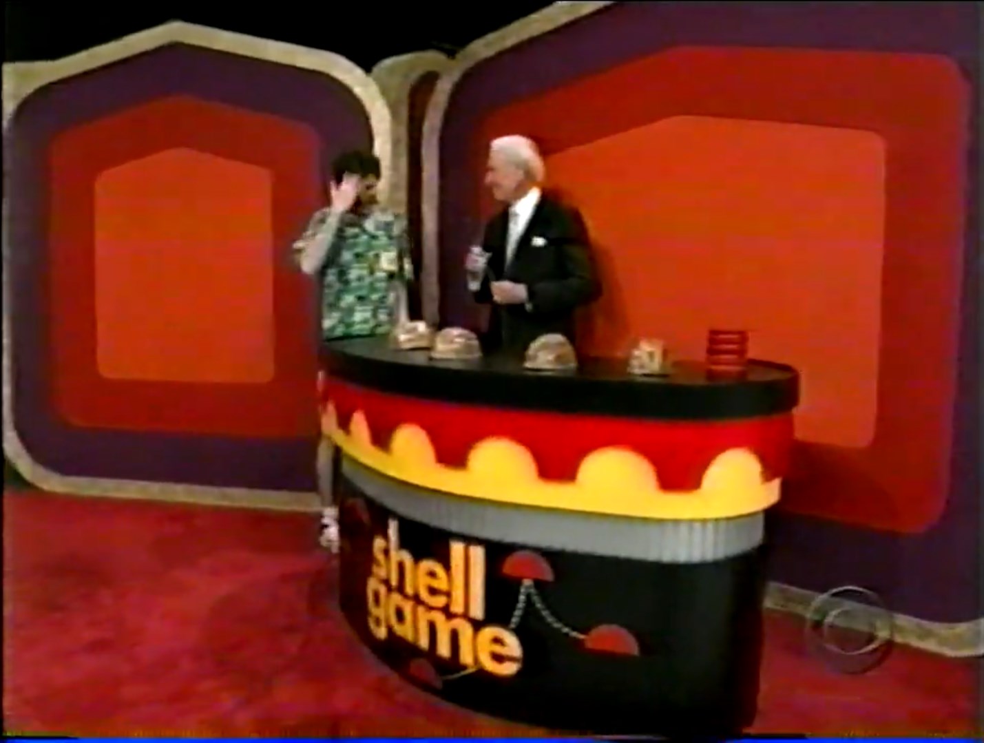 A contestant and the host standing by the Shell Game setup with multiple shells on a table and a colorful backdrop.